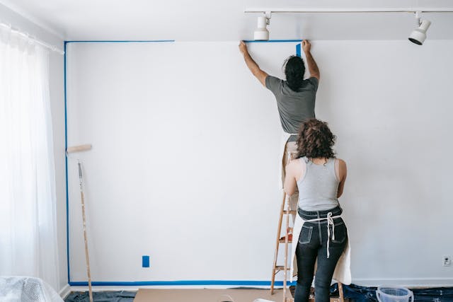 A man painting a wall while a woman stands behind him holding a ladder