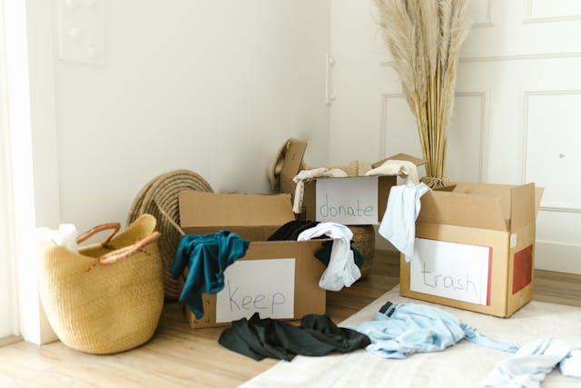 Boxes with labels for ‘keep’, ‘donate’, and ‘trash’ next to a pile of clothes on the floor