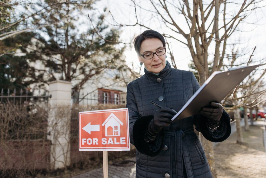 Woman in a jacket holding notes next to a house for sale banner